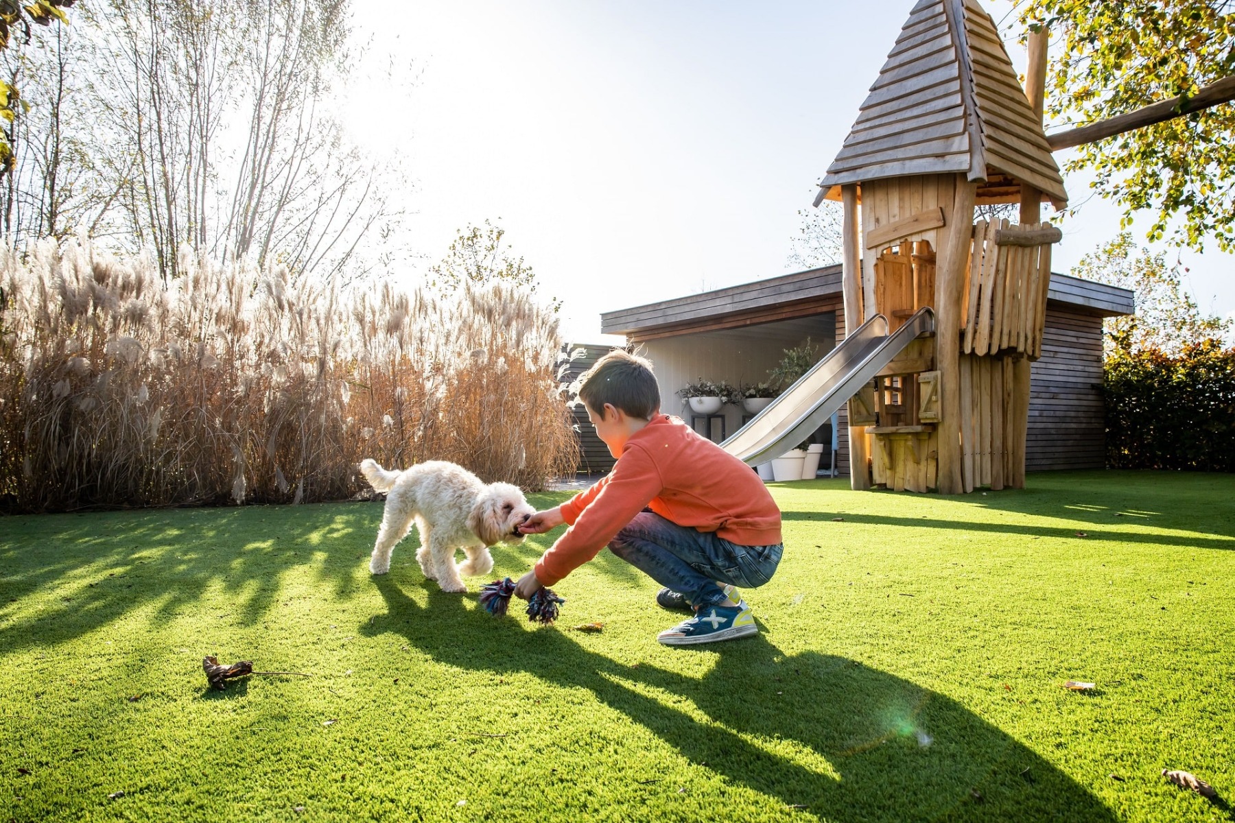 Een kind dat samen met zijn hond in de tuin op een kunstgras mat speelt. Op de achtergrond schijnt de zon en staan een tuinhuis en speelhut met glijbaan afgebeeld.